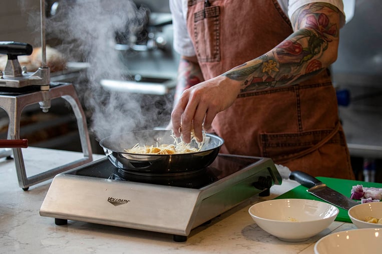 chef sprinkling shredded cheese in a pan of food cooking on an induction stove